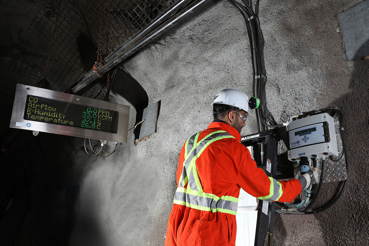 Miner in an underground tunnel, attaching the Maestro Digital Mine Vigilante AQS to their system. A Superbrite Display unit is in the background hanging from the tunnel roof.